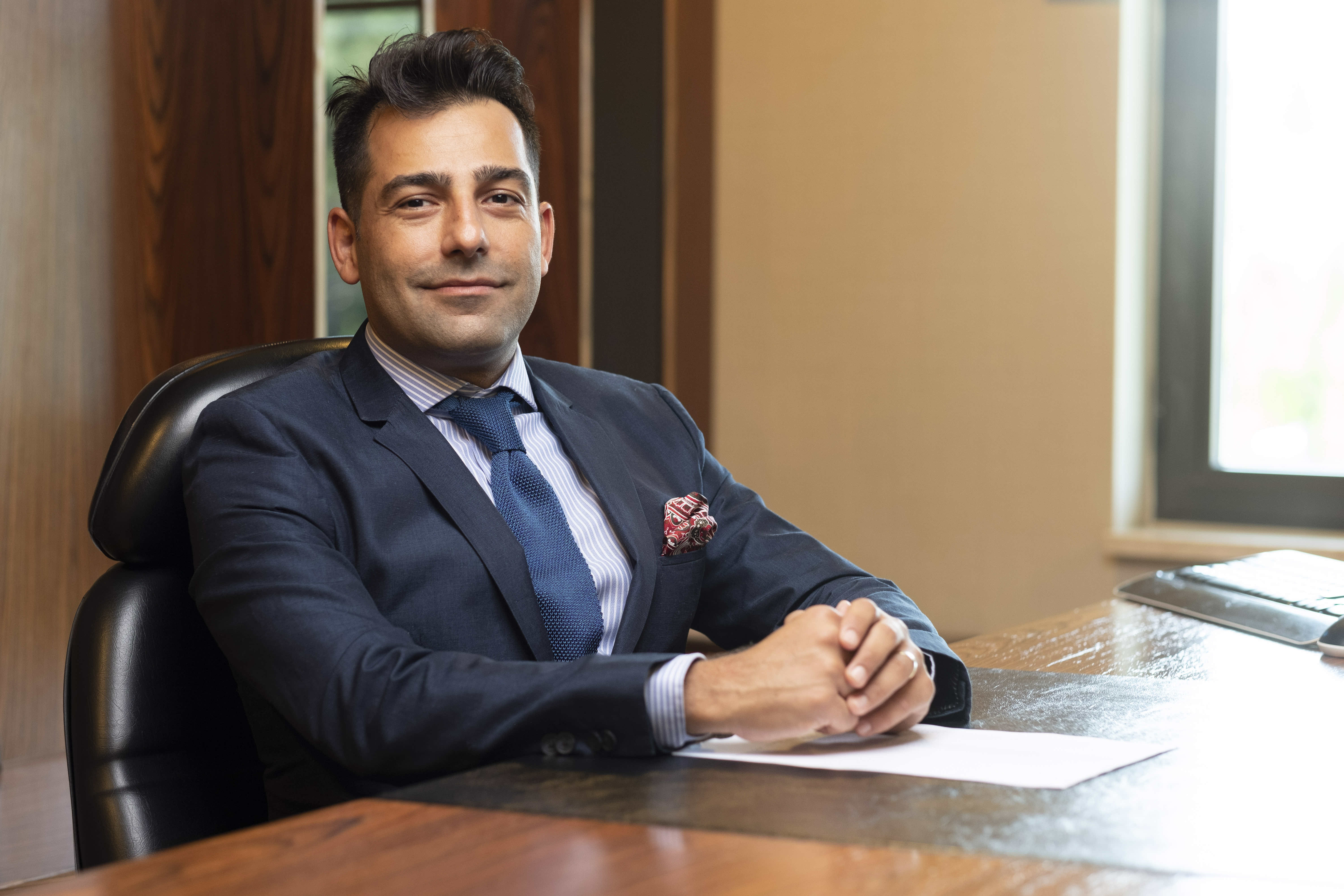 Professional business portrait of Ezgican Aydoğan in a navy blue suit with blue tie and decorative pocket square, seated confidently at a marble desk in an elegant office setting with warm lighting and modern professional background